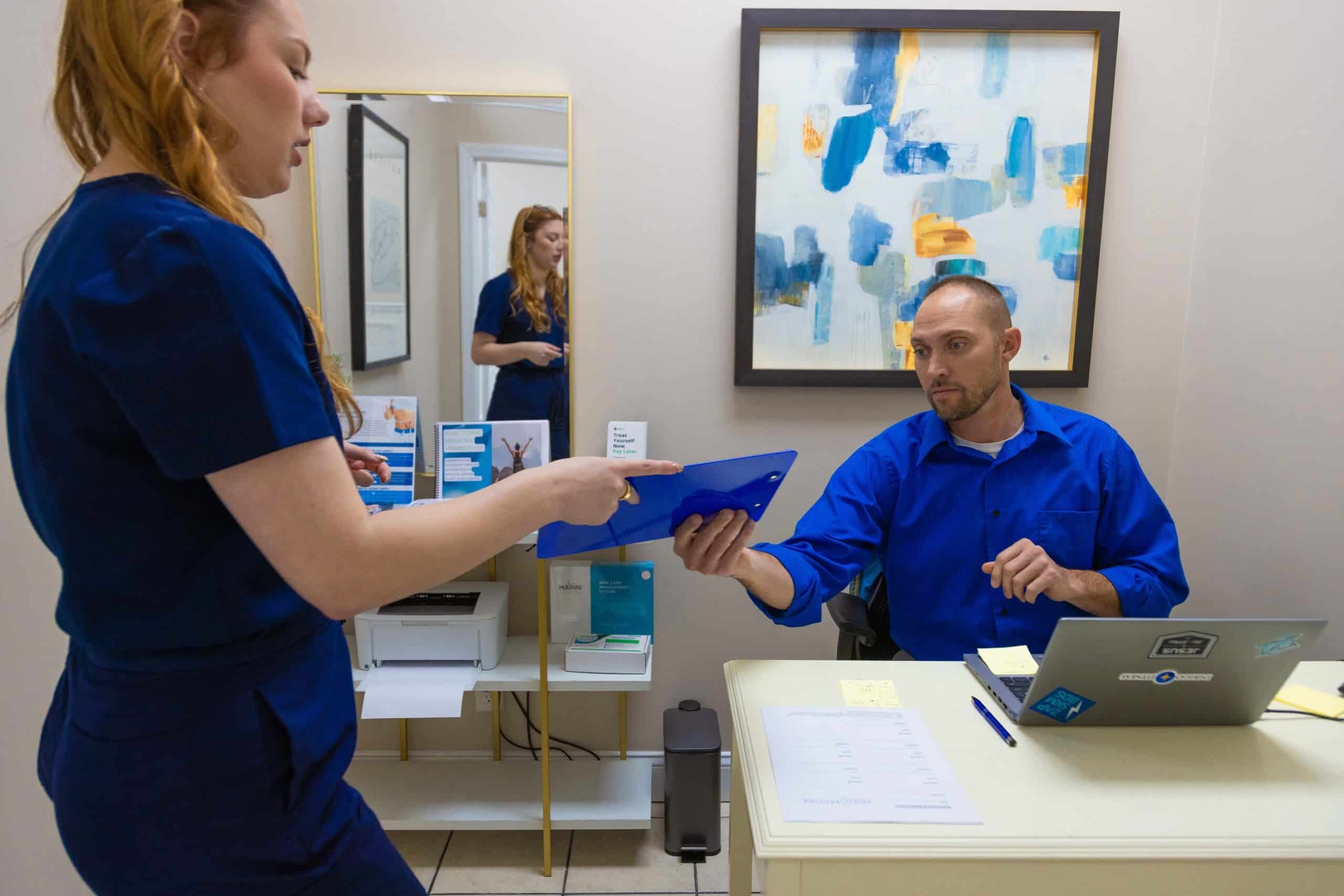 Woman in navy scrubs handing a blue clipboard to a man seated at a desk with a laptop