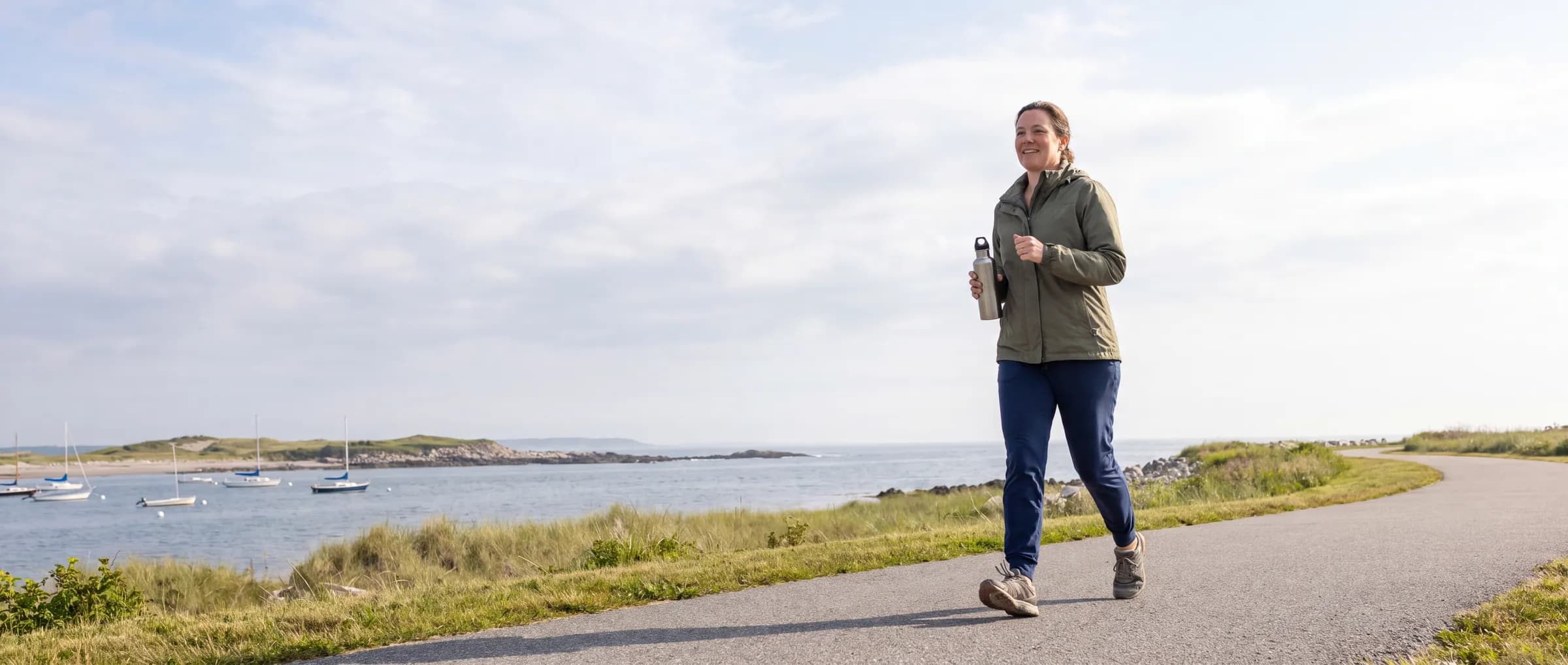 A woman walking a paved path beside the water, carrying a bottle