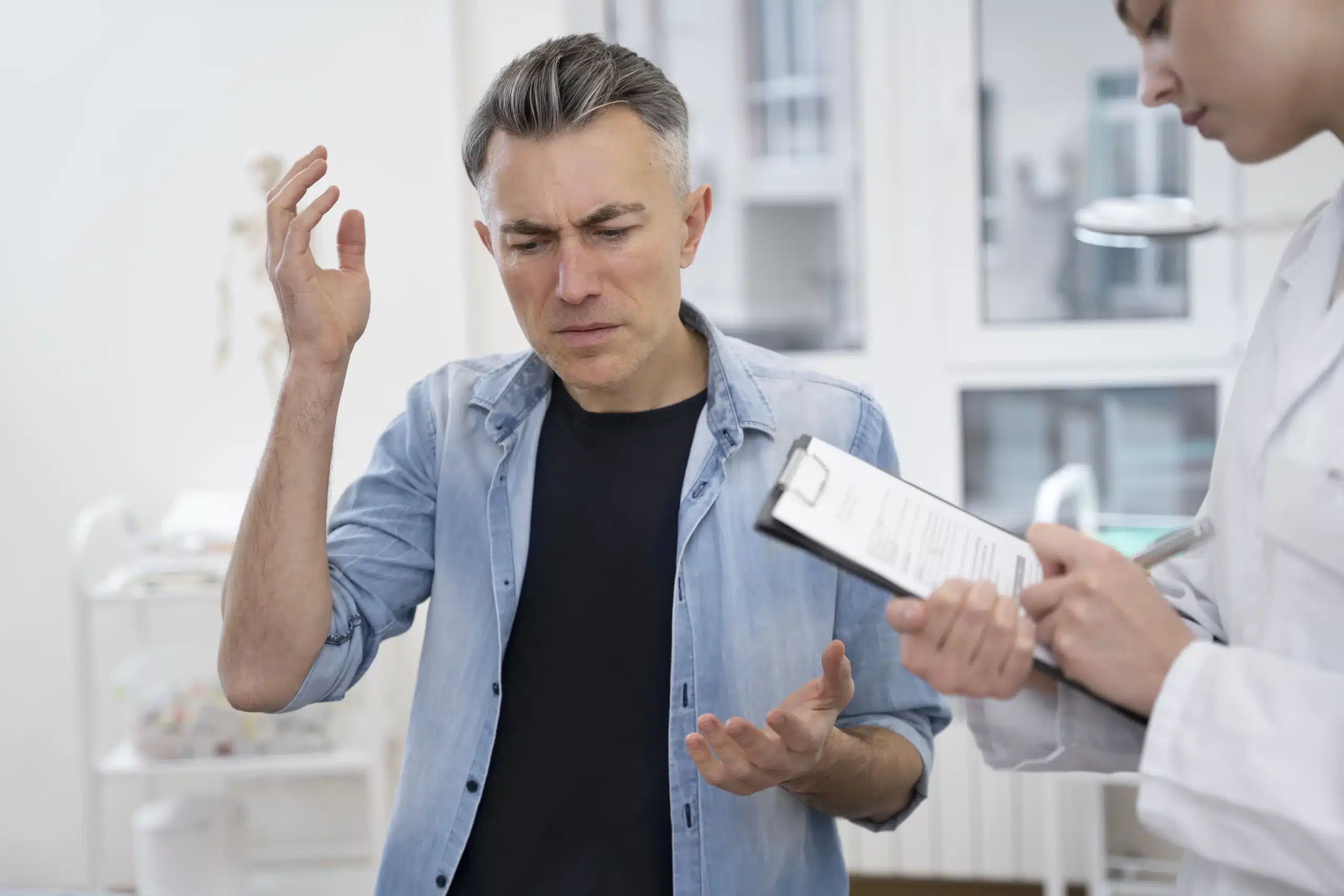 A man appears to be describing his symptoms with a pained expression while a healthcare professional takes notes on a clipboard, discussing affordable hormone therapy options. in Portland Maine