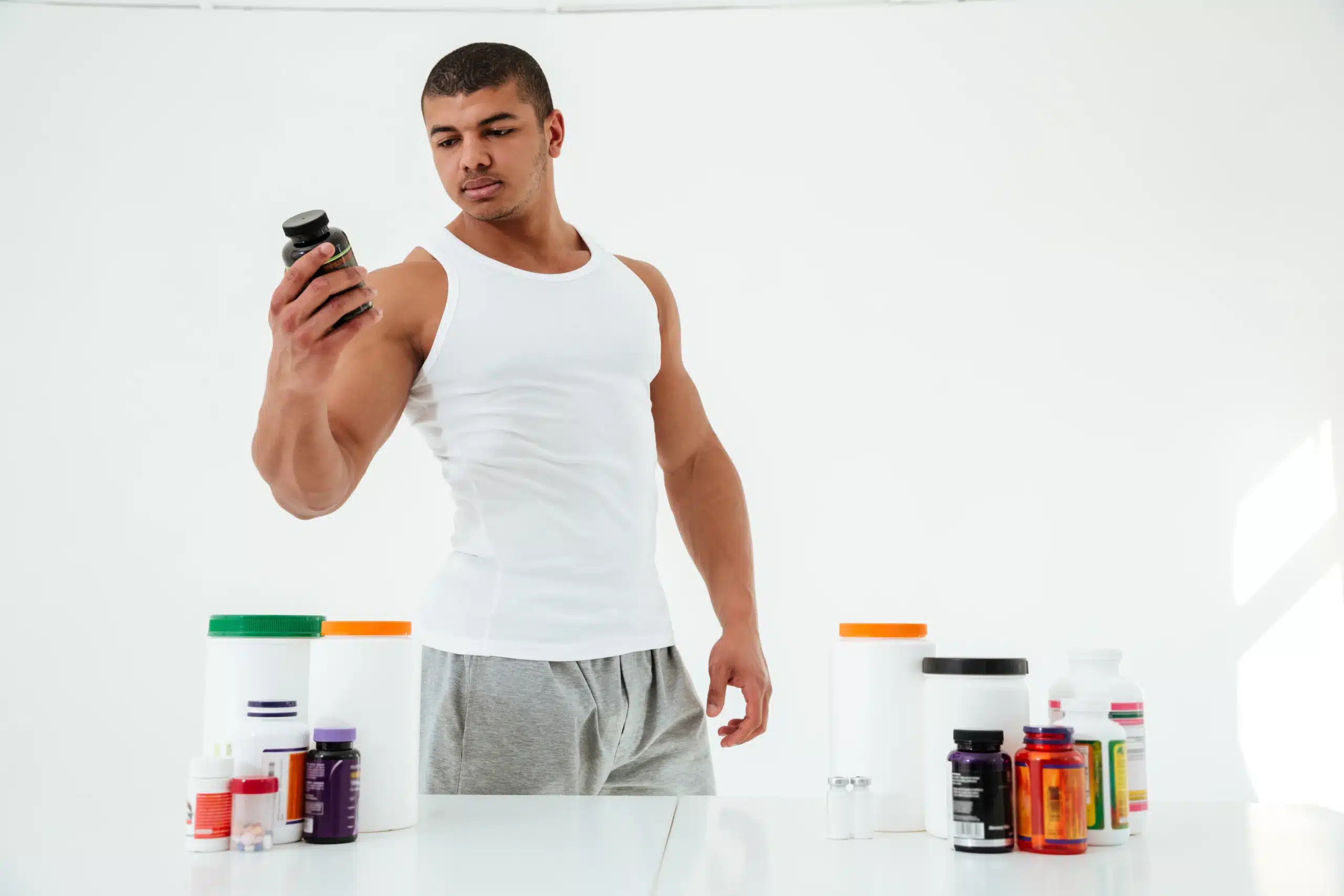 A man in a white tank top examines a supplement bottle while standing at a table with various supplement containers, highlighting the focus of integrative medicine for athletes. in Portland Maine