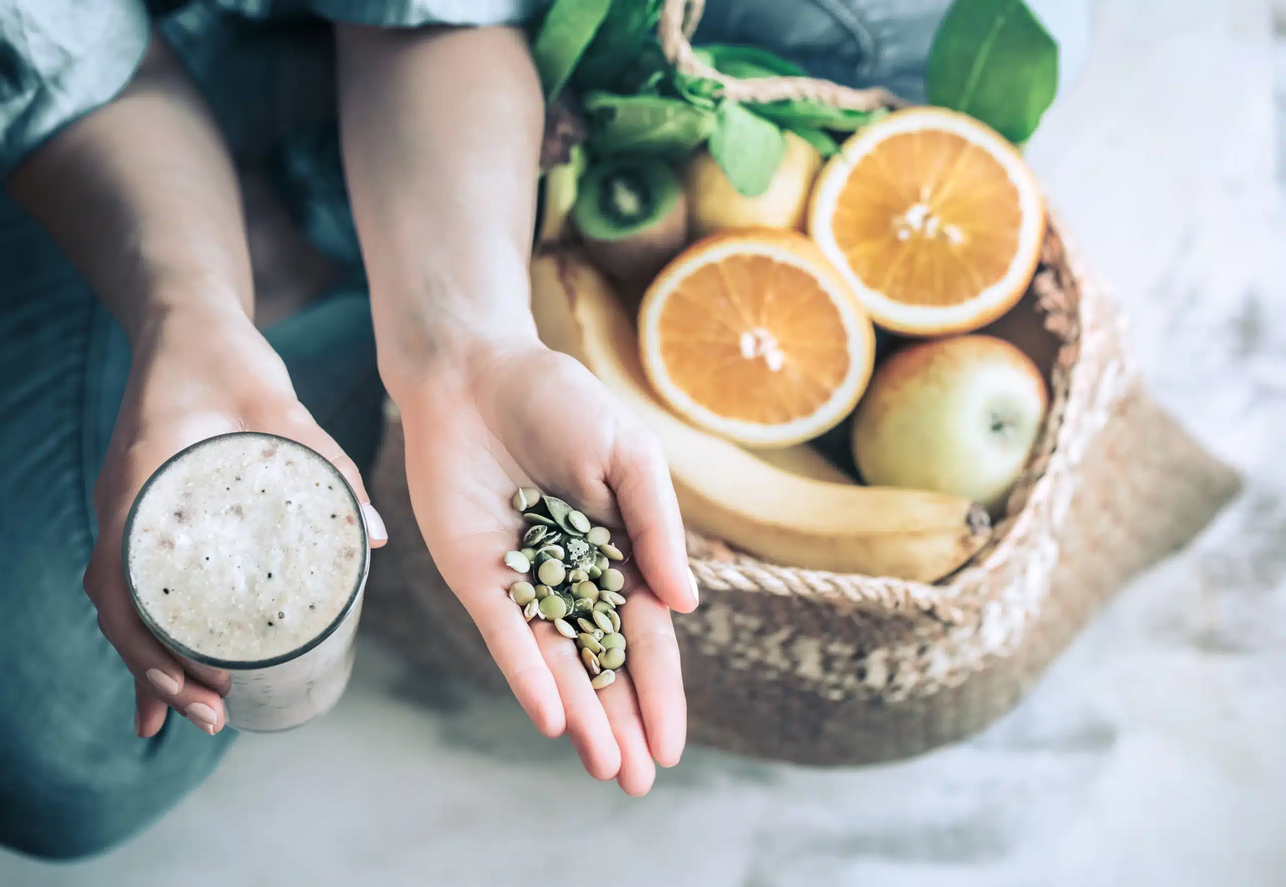 Person holding a glass of smoothie and a handful of seeds, next to a basket containing oranges, bananas, apples, kiwi, and leafy greens—perfect ingredients for holistic detox approaches. in Portland Maine