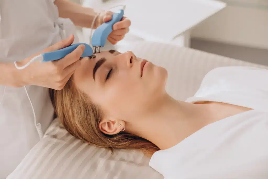 Person lying on a treatment bed receiving a facial procedure with two handheld blue devices applied to the forehead by a professional at an ozone therapy clinic. in Portland Maine