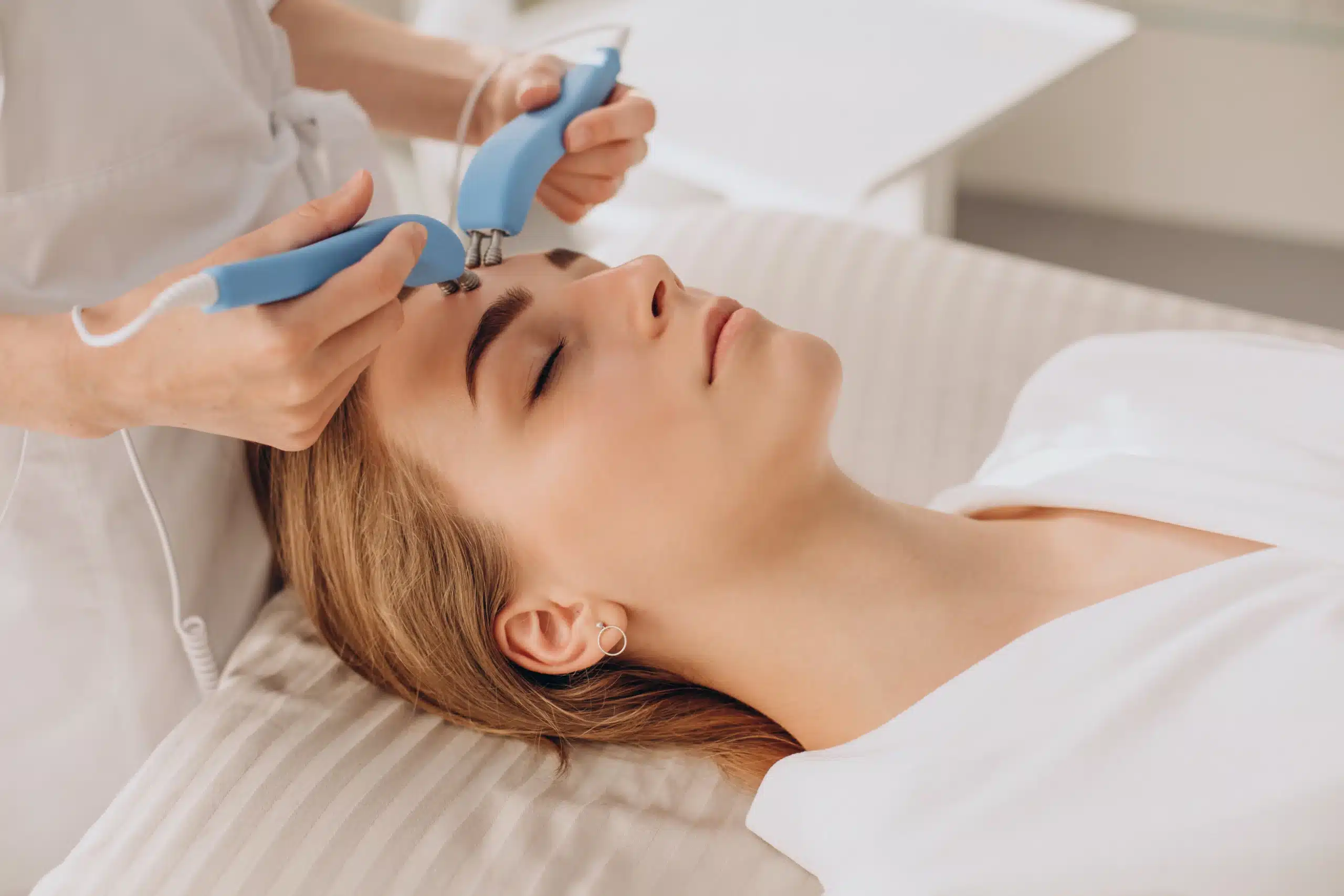 Person lying on a treatment bed receiving a facial procedure with two handheld blue devices applied to the forehead by a professional at an ozone therapy clinic. in Portland Maine