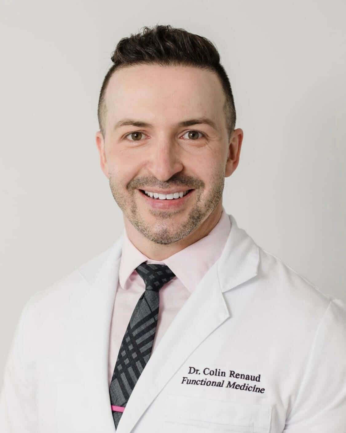 A man wearing a white lab coat, dress shirt, and patterned tie, smiling at the camera. The lab coat reads "Dr. Colin Renaud, Functional Medicine," reflecting his commitment to the team's mission of better health.