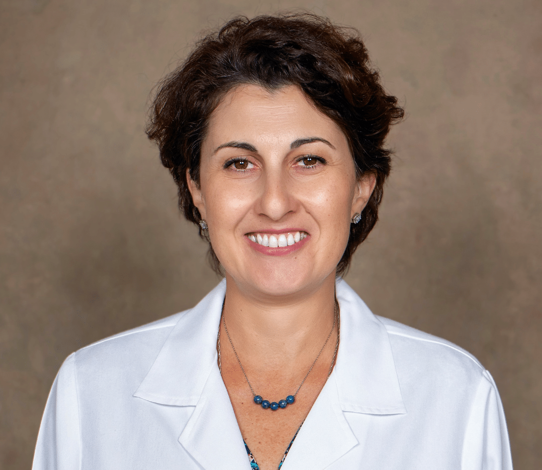 Sophia Viner, DNP, ANP-BC, with short brown hair, smiles at the camera. She is wearing a white lab coat and a blue beaded necklace, standing against a plain brown background.